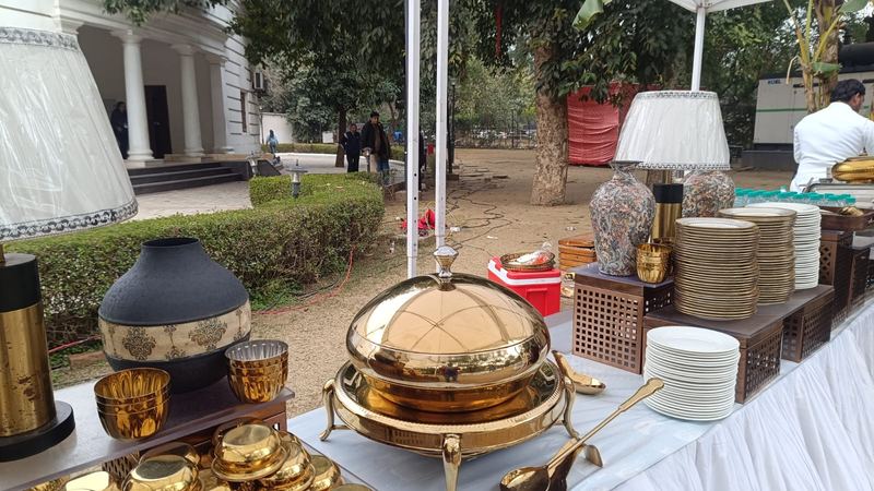 Outdoor wedding buffet with a polished brass chafing dish, gold-rimmed plates and decorative urns