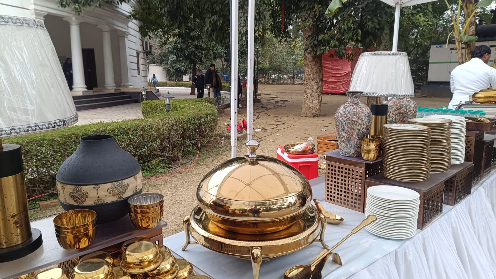 Outdoor wedding buffet with a polished brass chafing dish, gold-rimmed plates and decorative urns